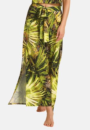 Kobieta w zielonej spódnicy z tropikalnym liściastym i lampartim wzorem, z rozporkiem z boku, boso na jednolitym białym tle.