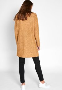 Lång, ribbad tröja i senapsgul, tillverkad av en texturerad stickning. Kombinerad med svarta tighta byxor och vita sneakers. Baksidan visas.