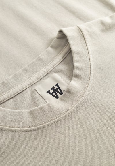 Ανοιχτό μπεζ T-shirt με στρογγυλή λαιμόκοψη, απαλή υφή υφάσματος και ετικέτα με λογότυπο ραμμένη στον λαιμό.