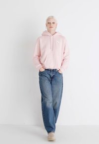 Ljusrosa hoodie med ficka fram, kombinerad med lösa blå jeans och beige skor, mot en vit bakgrund.