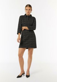 Schwarzes Kleid mit weißem Karomuster, mit Kragen, langen Ärmeln und schmalem Schnitt. Kombiniert mit schwarzen spitzen Absatzschuhen.