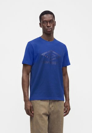 Νέος άνδρας που φορά ένα φωτεινό μπλε t-shirt με κοντά μανίκια και ένα σκούρο γεωμετρικό λογότυπο και ανοιχτό καφέ παντελόνι, στέκεται μπροστά σε ένα απλό φόντο.