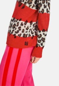 Rot gestreifter Pullover mit Leopardenmuster, getragen über rosa Hosen mit roten seitlichen Streifen. Auffälliger Textur- und Farbkontrast.