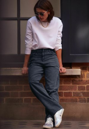 Gray corduroy pants, white long-sleeve shirt, silver chain, and round sunglasses. Worn with white sneakers, seated on a brick ledge.