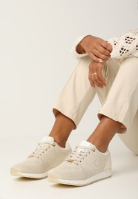 Beige stickade sneakers med texturerad ovandel, vita accenter och logodetalj. Matchas med ljusbeiga cropped byxor och en spetsig topp.