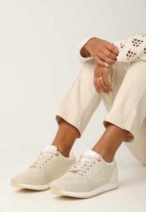 Beige gebreide sneakers met een gestructureerde bovenkant, witte accenten en logo-details. Gecombineerd met lichte beige cropped broek en een kanten top.