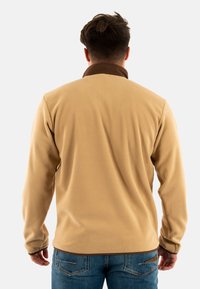 Chaqueta de forro polar beige con un collar marrón, mangas largas y una textura suave, vista desde atrás. Combinada con jeans de mezclilla azul.