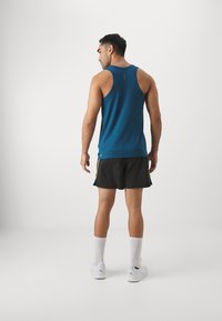 Camiseta sin mangas azul para hombre con un diseño de espalda nadadora, que presenta un patrón texturizado. Combinada con pantalones cortos negros con detalles verdes y zapatillas blancas.
