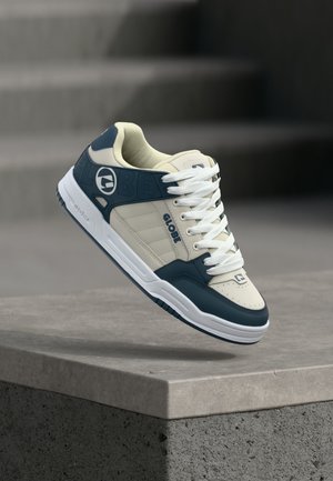 Beige en blauwe Globe merk sneaker met witte veters zwevend boven een grijs betonnen rand met trappen vaag op de achtergrond.