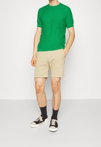 Grön texturerad kortärmad tröja i kombination med beiga shorts, svarta strumpor och svarta sneakers, vilket visar en avslappnad sommaroutfit.