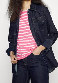 Osoba v tmavě modré džínové bundě a kalhotách s růžovým a bílým pruhovaným tričkem, ruka v kapse, záběr od ramen po stehna.