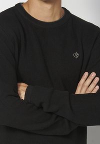 Černý žebrovaný svetr s kulatým výstřihem, zdobený malým vyšitým logem na hrudi. Měkká textura a volný střih.