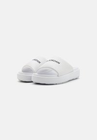 White slide sandals with a wide strap and embossed "JORDAN" logo. The sole is thick, textured for grip, and slightly raised at the heel.