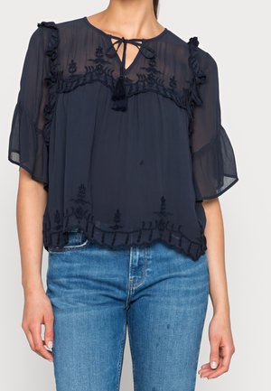 Blusa trasparente blu navy con bordo in pizzo, maniche arricciate, scollo con fiocco e ricamo floreale lungo l'orlo e le spalle. Abbinata a jeans.