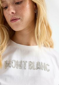 Vit bomulls T-shirt med "MONT BLANC" i glittrande rhinstenar. Långt blont hår och hängande rhinstenörhängen syns.