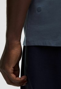 T-shirt bleu à manches courtes en tissu doux, avec un petit logo sur l'ourlet et une coupe décontractée, associé à un pantalon foncé.