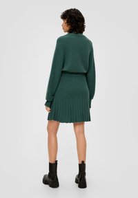 Groene gebreide jurk met ribbelstructuur, aansluitende lijfje en geplooide rok. Lange mouwen met schouderaccenten. Zwarte enkelboots.