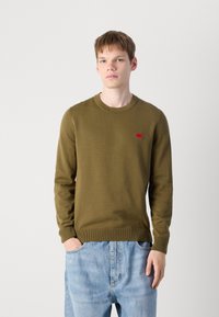 Pull olive vert avec un col rond, des poignets et un ourlet côtelés. Présente un petit logo rouge sur la poitrine. Associé à un jean bleu clair.