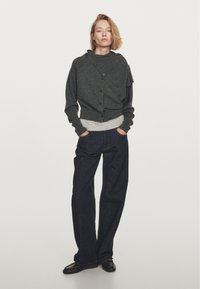 Grijze, textuur cardigan met een capuchon en knoopsluiting, gelaagd over een lichtgrijs shirt. Draagt het met losse, donkerdenim jeans en zwarte schoenen.