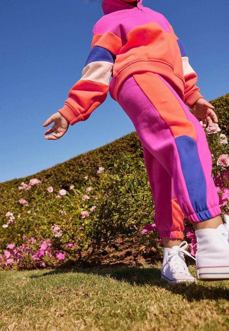 Enfant portant un survêtement coloré rose, orange et violet ainsi que des baskets montantes blanches, marchant près de buissons en fleurs sous un ciel bleu clair.