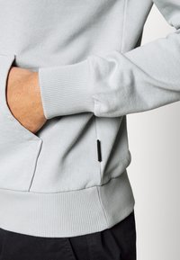 Main dans la poche d'un sweat-shirt gris clair, montrant le poignet et l'ourlet côtelés, avec un pantalon noir visible en dessous.
