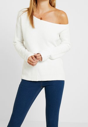 Witte off-shoulder ribbed sweater met lange mouwen, voorzien van een gestructureerd breipatroon en een rechte onderrand, gedragen met blauwe jeans.