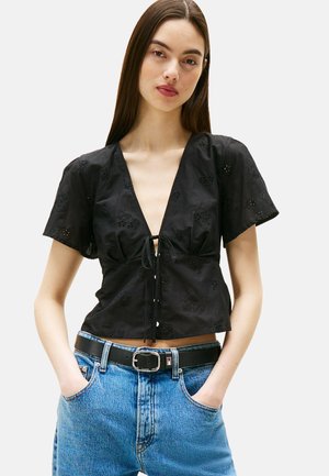 Schwarzes kurzärmeliges Oberteil mit tiefem V-Ausschnitt und Schleifenverschluss, verziert mit floralem Stickerei. Kombiniert mit hochtaillierten blauen Denim-Jeans und einem Gürtel.
