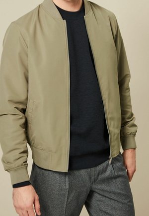 Homme portant une veste bomber marron clair sur une chemise noire texturée et un pantalon gris à carreaux sur un fond beige.