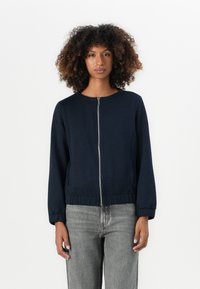 Junge Person mit lockigem Haar, die eine dunkelblaue Reißverschlussjacke und hellgraue Jeans trägt, steht vor einem schlichten weißen Hintergrund.