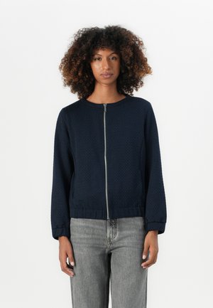 Junge Person mit lockigem Haar, die eine dunkelblaue Reißverschlussjacke und hellgraue Jeans trägt, steht vor einem schlichten weißen Hintergrund.