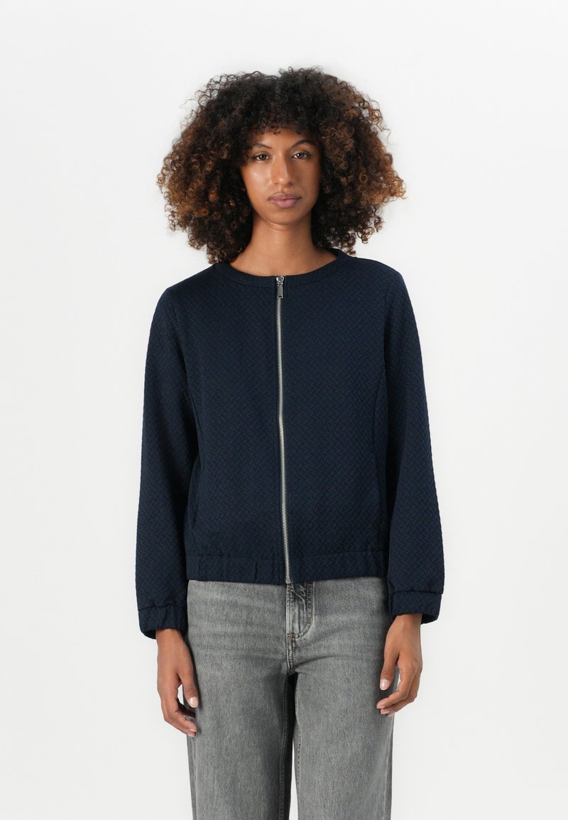 Junge Person mit lockigem Haar, die eine dunkelblaue Reißverschlussjacke und hellgraue Jeans trägt, steht vor einem schlichten weißen Hintergrund.