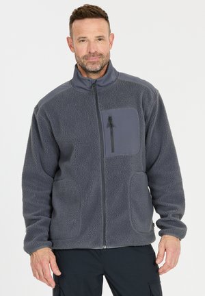 Mand iført mørkegrå fleecejakke med lynlåslomme på brystet og sorte bukser, stående foran en ensfarvet lys baggrund.