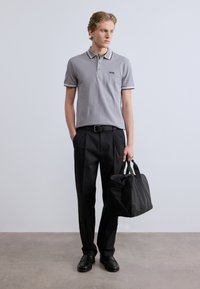 Ανοιχτό γκρι πόλο μπλουζάκι με μαύρες ρίγες στο γιακά, συνδυασμένο με μαύρο παντελόνι. Το μοντέλο κρατά μια μαύρη τσάντα με κομψό σχεδιασμό.