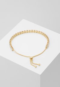 Zelta aproce ar kvadrātveida posmiem ar teksturētu apdari, regulējamu ķēdi un mazām apaļām pērlītēm, akcentēta ar caurspīdīgiem kristāliem.