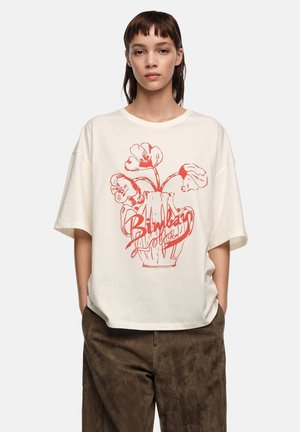 Persona joven con camiseta oversize color crema con gráfico de jarrón floral rojo y texto, combinada con pantalones marrones holgados, manos en los bolsillos.