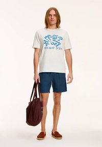Tricou alb cu text grafic albastru „HOLIDAY OFTEN”, pantaloni scurți bleumarin, pantofi maro slip-on și geantă tote maro închis. Ținută casual de vară.