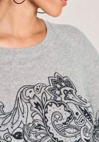 Frau in grauem Pullover mit detailliertem schwarzen Paisley-Blütenmuster und großen Kreolen, gezeigt vom Hals bis zur oberen Brust.
