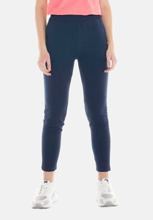 Personne portant un pantalon slim bleu marine, des baskets blanches et un haut rose, debout les mains détendues le long du corps.
