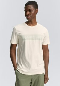 Camiseta de manga corta color crema hecha de una tela suave, con un logo impreso de "DENIM TOM TAILOR" en un rectángulo verde apagado en la parte frontal.
