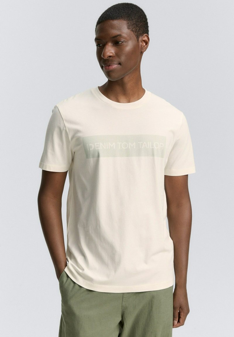 Camiseta de manga corta color crema hecha de una tela suave, con un logo impreso de "DENIM TOM TAILOR" en un rectángulo verde apagado en la parte frontal.