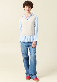 Hellgrau gestrickte Weste über einem hellblauen Hemd mit Knopfleiste, kombiniert mit weit geschnittenen blauen Jeans und roten Turnschuhen. Auffälliger V-Ausschnitt und gerippter Saum.