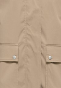 Beiger Stoff mit zwei Klappentaschen, die durch silberne Druckknöpfe gesichert sind, zentrierte vertikale Naht.
