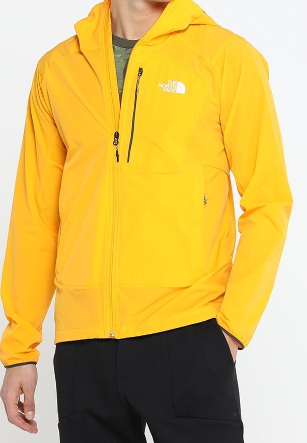 Homme portant une veste à capuche jaune vif avec des poches zippées à l'avant et sur la poitrine, associée à un pantalon noir et un t-shirt camouflage vert en dessous.
