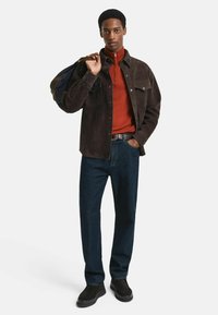 Brun mockajacka över en röd zip-tröja, ihop med mörkblå jeans och svarta skor; håller en blå väska. Enkel, modern design.