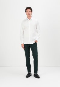 Weißes Hemd mit Knopfleiste, grüne Hose und schwarze Schuhe. Das Hemd hat lange Ärmel, während die Hose einen schmalen Schnitt und eine glatte Textur aufweist.