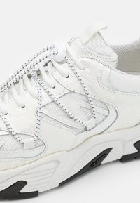 Vita sneakers med en texturerad läderöverkänga, mesh-paneler, kontrasterande svart gummi sula, och elastiska snören med vit låsknapp.