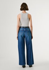 Weite blaue Jeans aus gewaschenem Baumwollstoff mit hoher Taille, zwei Gesäßtaschen und einem schwarzen Nieten-Gürtel. Graues Tanktop sichtbar.