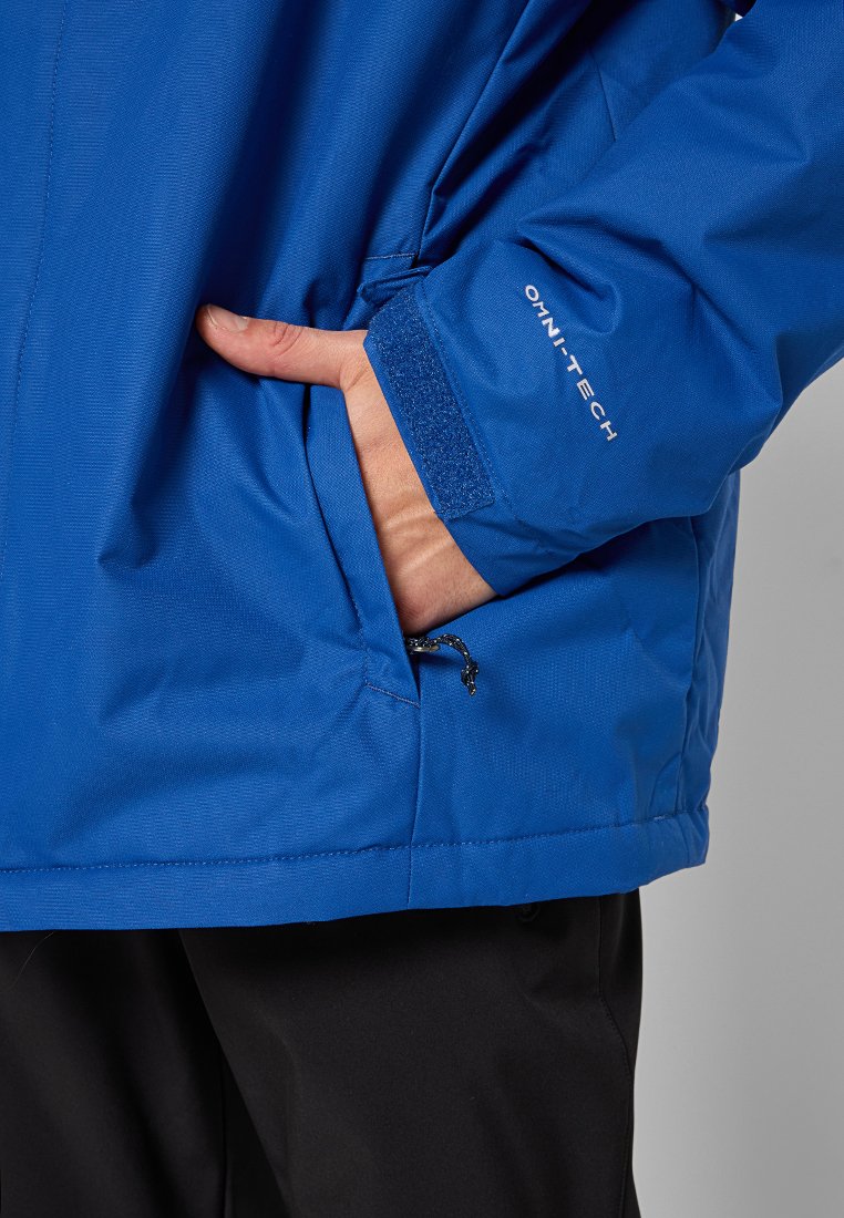 Blauwe waterdichte jas van gestructureerd materiaal, met een ritszak en klittenbandsluiting bij de manchetten. Logo zichtbaar op de mouw.