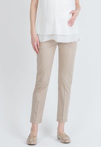 Pantalons beiges sur mesure en tissu lisse, à coupe slim et longueur cheville. Portés avec un blouse blanche à couches et des chaussures à motifs.