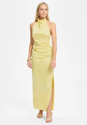 Femme portant une robe jaune en satin sans manches avec un col licou et une fente latérale, des sandales dorées, des boucles d'oreilles anneaux et un bracelet, debout devant un fond blanc.